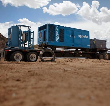 A large, dark blue containerized industrial unit is secured on a blue flatbed trailer in a dry, dirt field under a partly cloudy sky. 