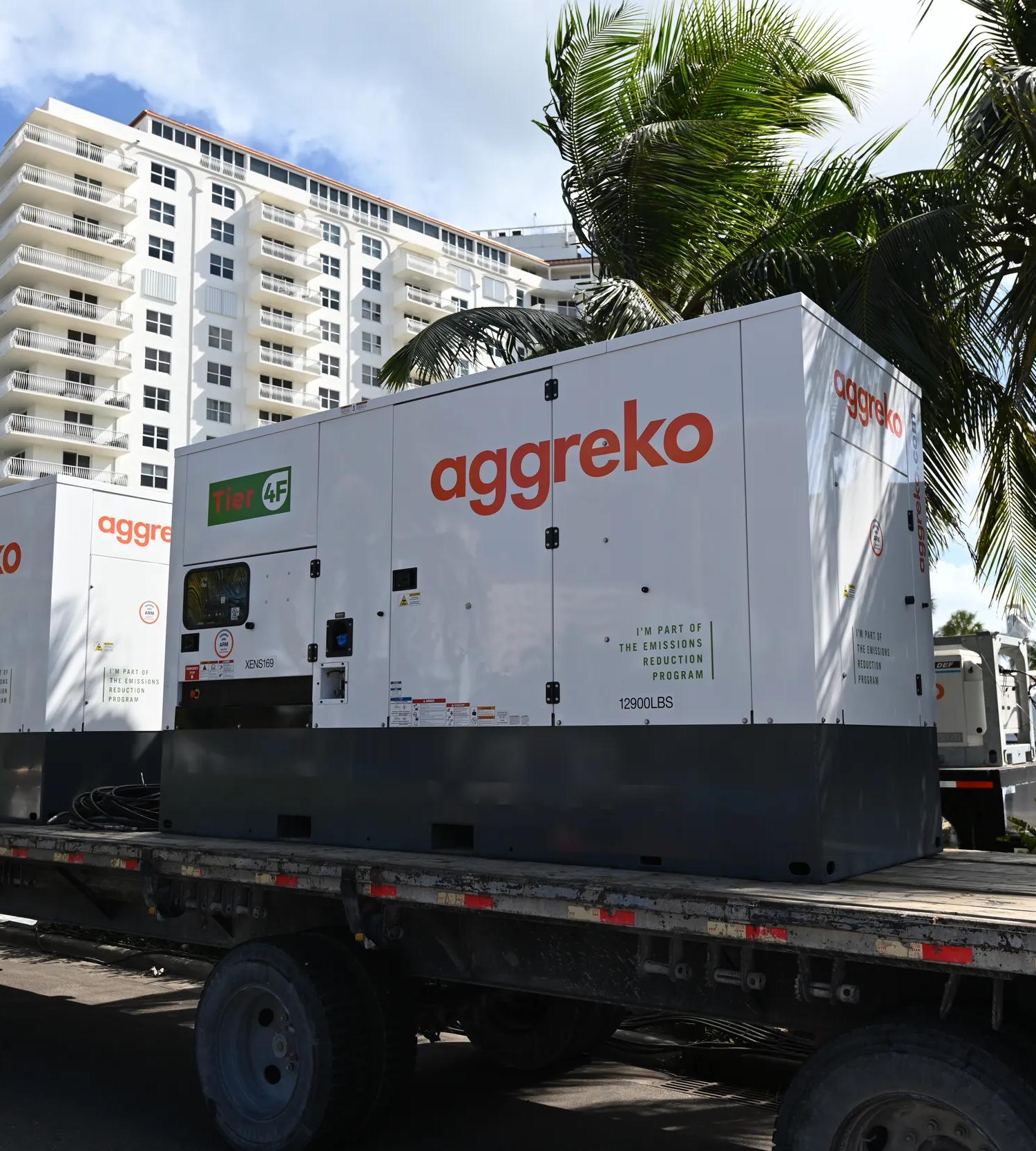 View of a large, white Aggreko generator outdoors, beside a palm tree.