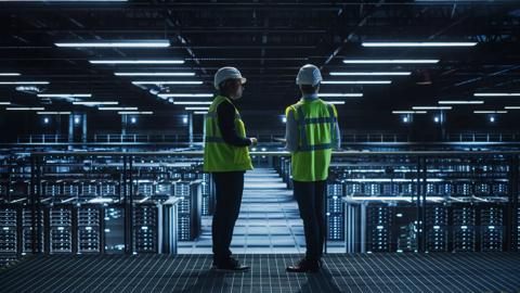 Data Center IT Specialist and System administrator Talk, Use Tablet Computer, Wearing Safety West. Server Cloud Farm Facility with Two Information Technology Engineers checking Cyber Security.