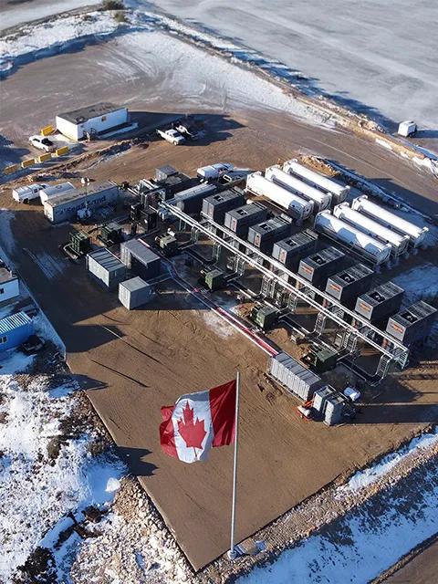 High-angle view of an industrial energy site in a snowy landscape. A large Canadian flag waves in the foreground, with rows of modular power generators behind it.