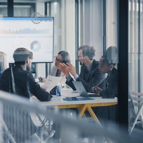 Business Leaders Sit Around a Conference Table Reviewing Charts on a Large Screen, Debating Results, Setting Priorities, and Planning Next Steps While Using Laptops, Printed Reports, and Notes During
