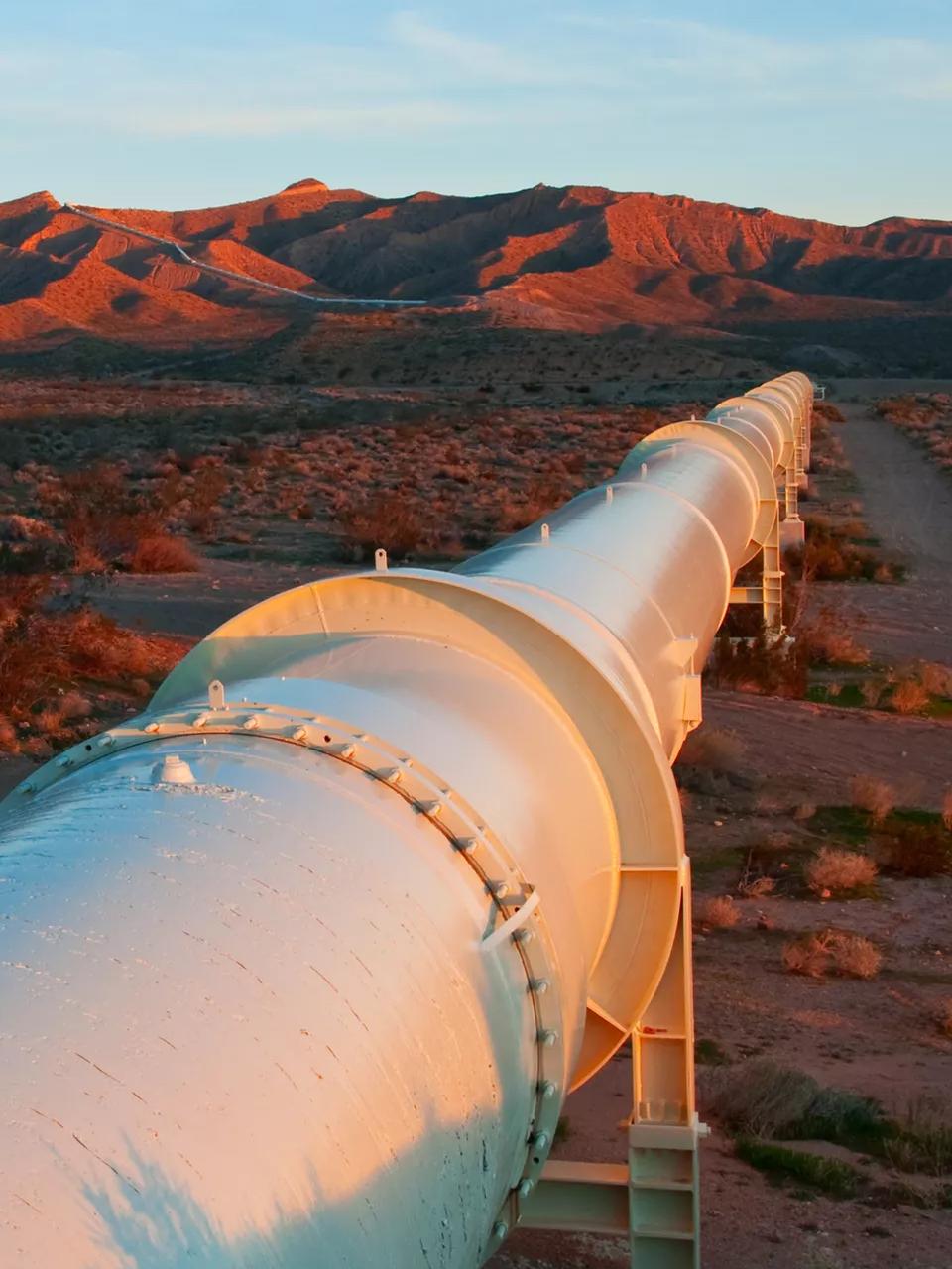 A low-angle view of a large, white industrial pipeline stretching across a dry, brush-filled desert landscape.