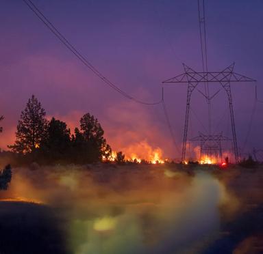 A high-voltage power transmission tower silhouetted against a red and purple wildfire sky.