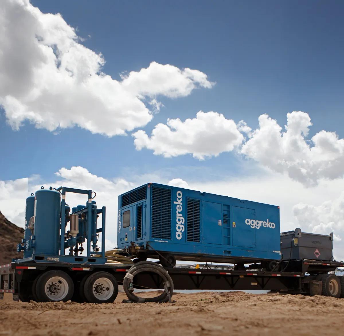 A dark blue, containerized industrial unit, labeled Aggreko, is secured on a flatbed trailer in a sunny, dry, dirt field.