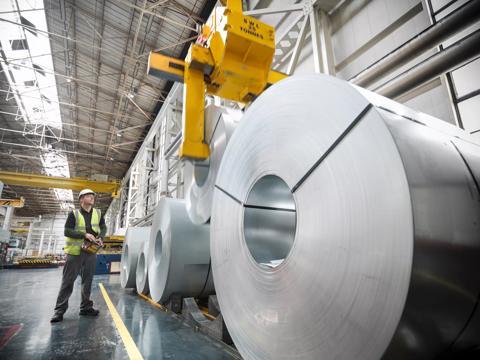 A worker in a safety vest supervises a crane lifting large rolls of metal in a spacious industrial warehouse.