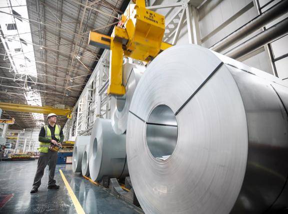 A worker in a safety vest supervises a crane lifting large rolls of metal in a spacious industrial warehouse.