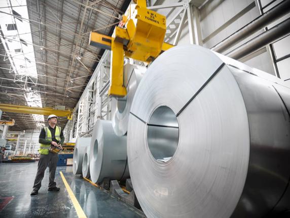 A worker in a safety vest supervises a crane lifting large rolls of metal in a spacious industrial warehouse.