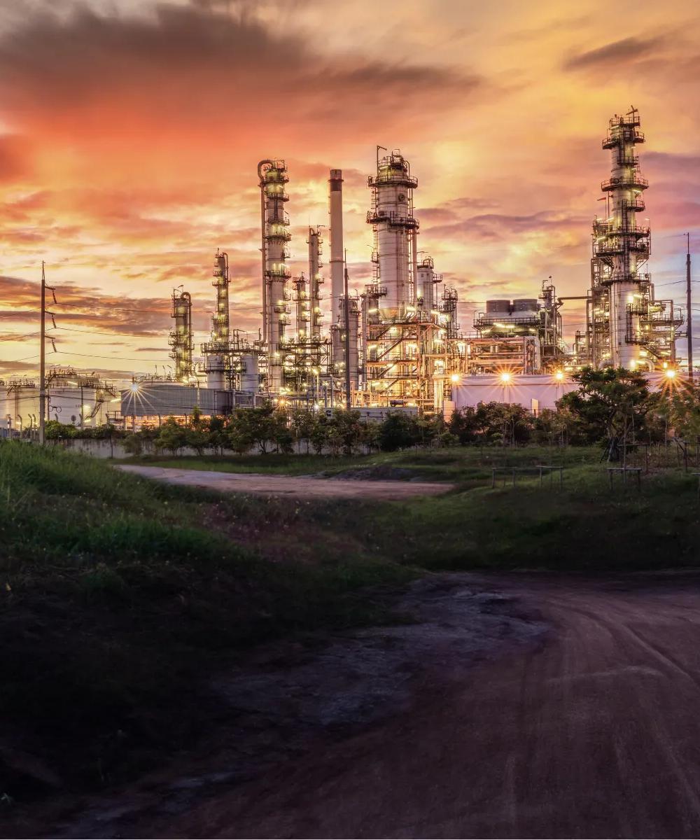 Vue d’une usine pétrochimique au bout d’un chemin de terre, avec en arrière-plan un ciel nuageux au coucher du soleil.