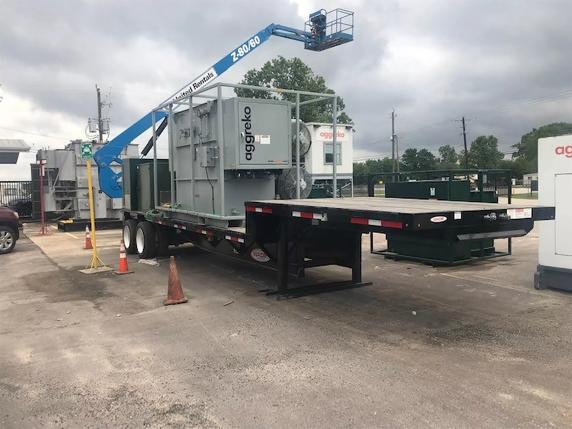 A large, gray, containerized Aggreko industrial unit is mounted on a heavy-duty flatbed trailer. A bright blue articulated boom lift is positioned next to the unit.