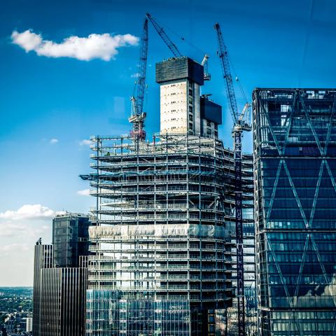 Low angle color image depicting a construction site in London, UK. A housing development is in the process of being created, and multiple cranes are hoisting materials into place. Beyond the cranes and construction equipment is a dazzling sky and cloudscape. Lots of room for copy space.