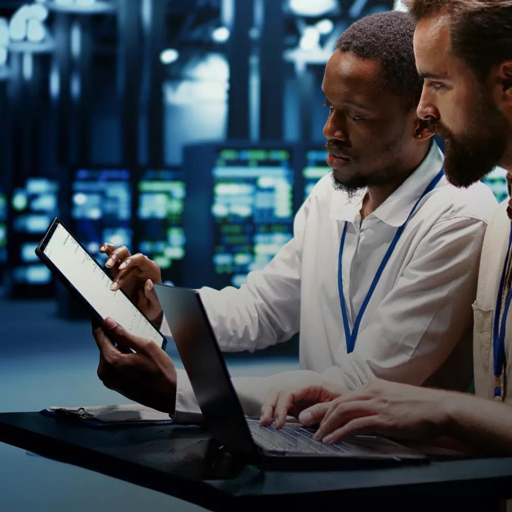 A man is holding a tablet and showing it to another man, who is typing on a laptop, inside a data center