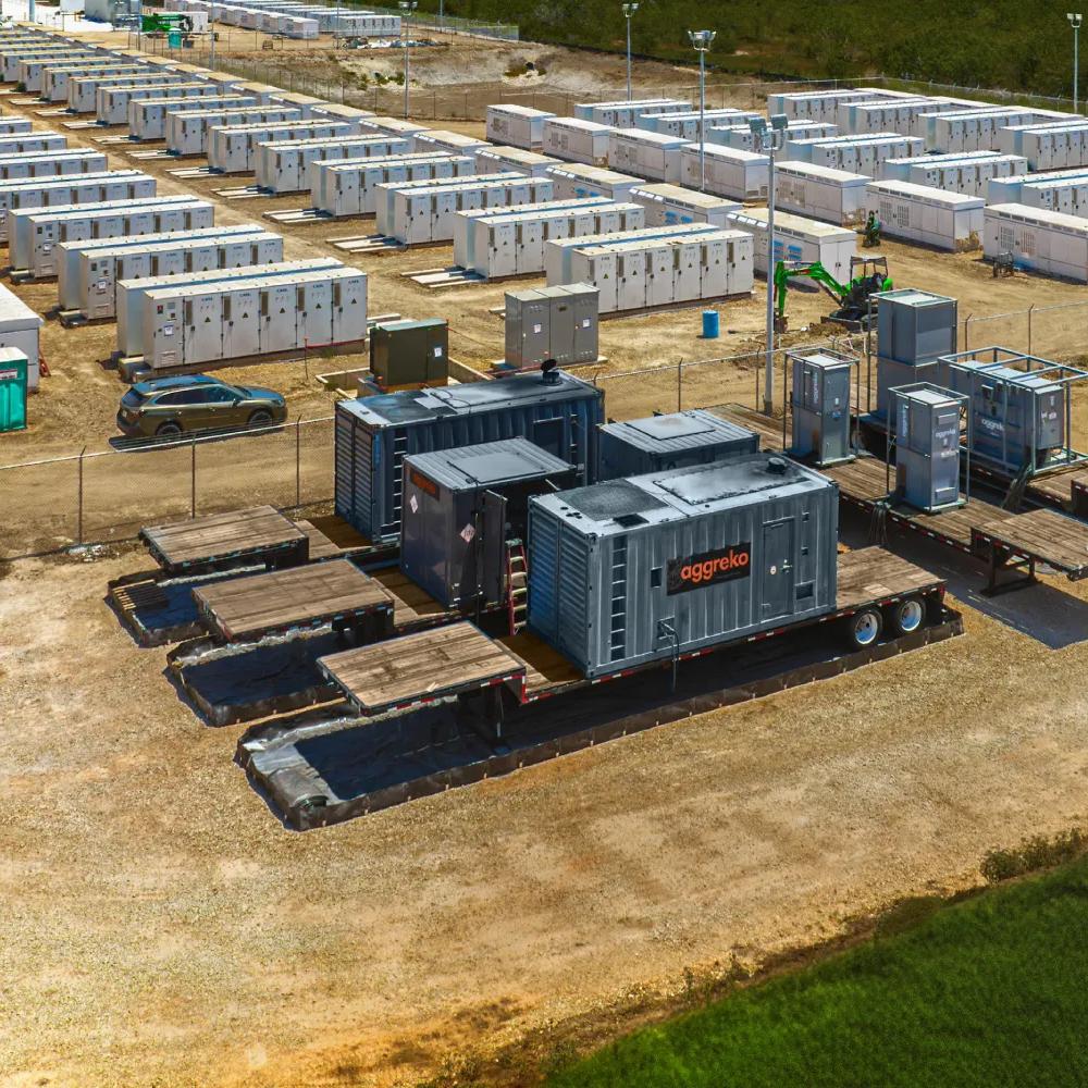 An aerial view of a a large, dark gray modular power unit  on a black trailer, with a vast, industrial staging area of hundreds of identical white containerized units arranged in rows in the background. In the foreground.