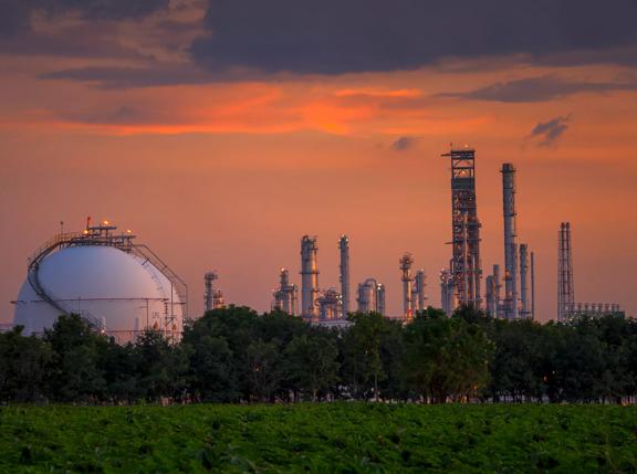  Industrial skyline at sunset featuring a gas storage dome and oil refinery towers, with greenery in the foreground.