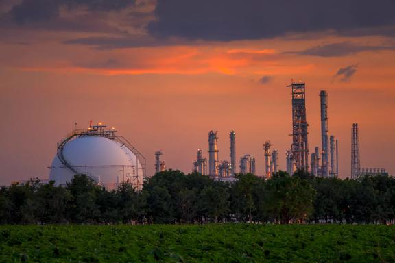  Industrial skyline at sunset featuring a gas storage dome and oil refinery towers, with greenery in the foreground.