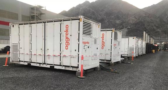 A perspective view of a row of large, white, containerized Aggreko industrial units installed on a gravel lot at the base of a steep, rocky mountain slope. 