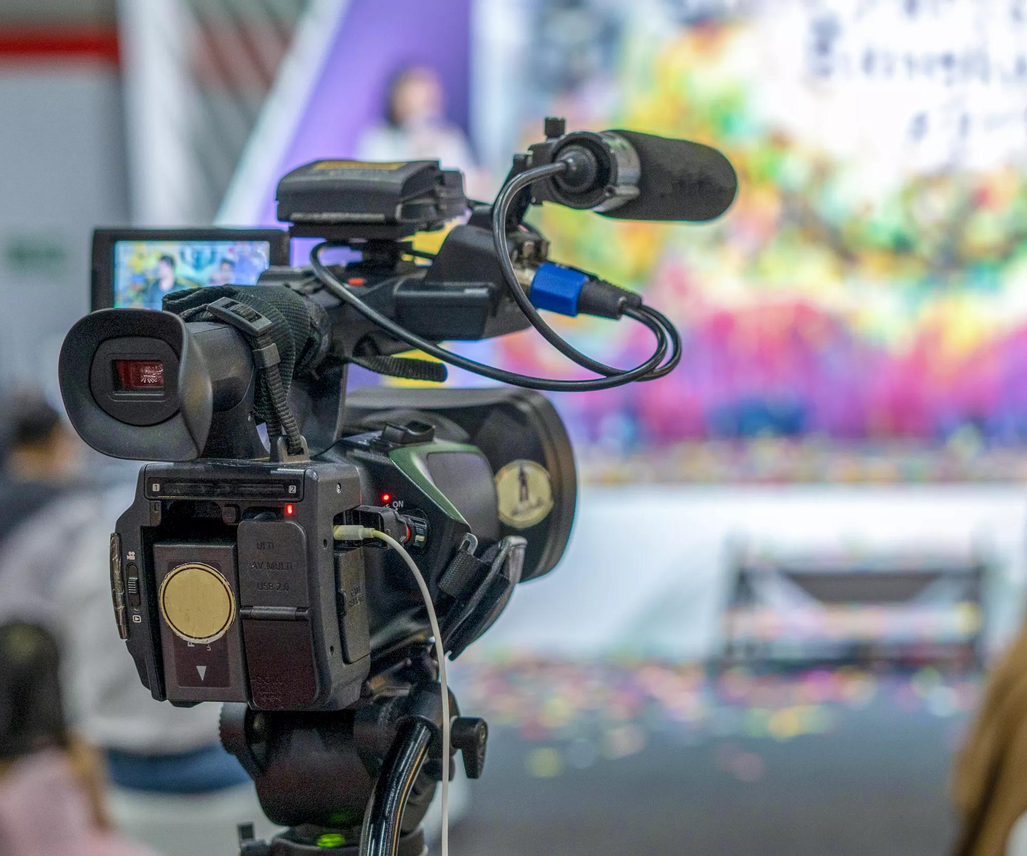 A close-up of a professional video camera pointing toward a blurry, colorful television production background.