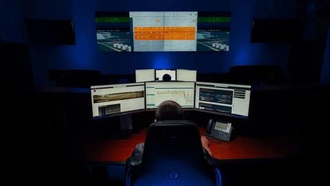 A dark control room with two workers seated at different desks with three computer monitors. Two screens in the background turned off. Two screens on the back, with a big central screen displaying performance metrics.