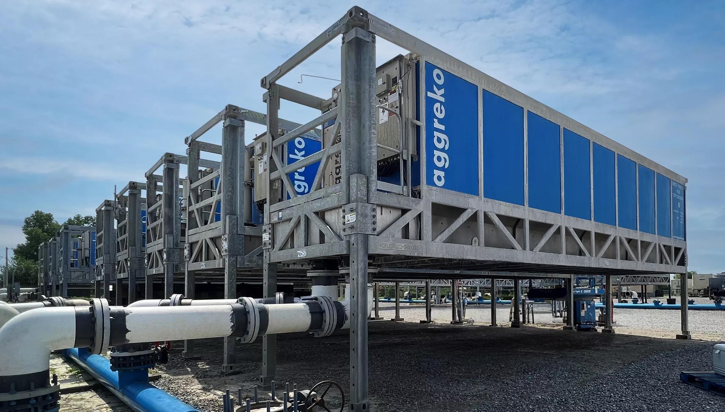 Row of elevated blue industrial container units with large white pipes in the foreground.