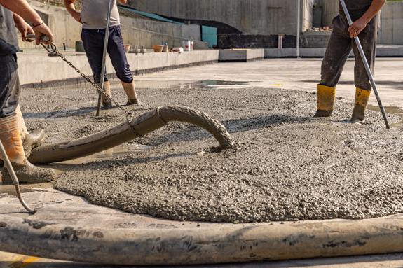 A close-up shot of a construction site where a thick, gray concrete mix is actively being poured onto a slab. Three construction workers wearing rubber boots are visible around the edges of the growing concrete pool.