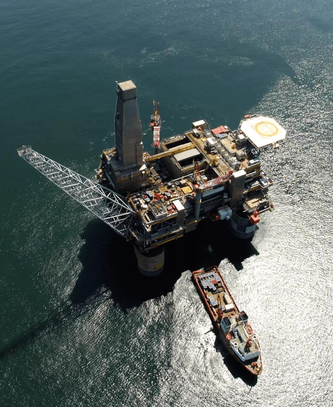 An aerial view looking straight down onto a large, complex offshore oil platform with a helicopter landing pad, surrounded by deep blue ocean water under bright sunlight.