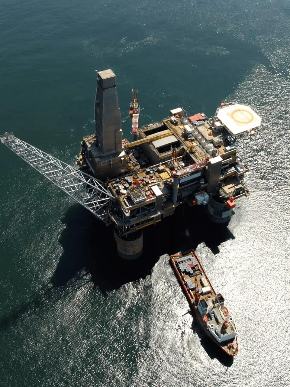 An aerial view looking straight down onto a large, complex offshore oil platform with a helicopter landing pad, surrounded by deep blue ocean water under bright sunlight.