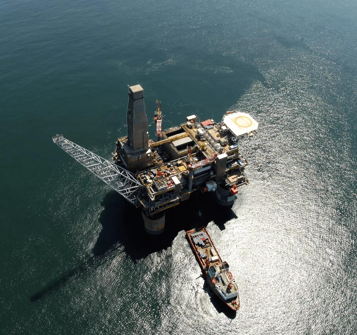 An aerial view looking straight down onto a large, complex offshore oil platform with a helicopter landing pad, surrounded by deep blue ocean water under bright sunlight.