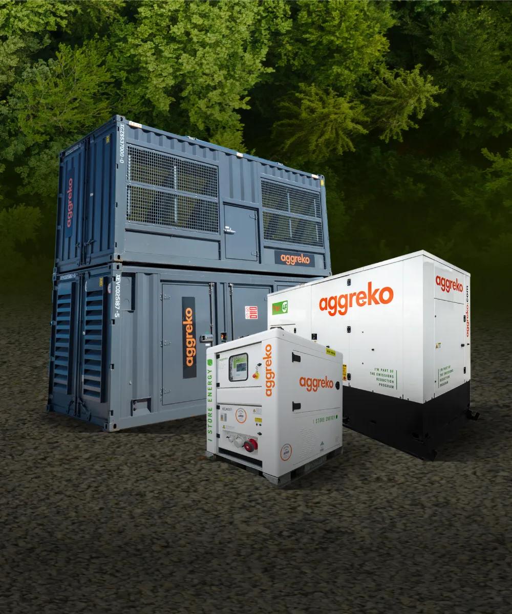 Three Aggreko power units, two white generators, and a large, stacked gray container unit, all on dirt in front of a forest.