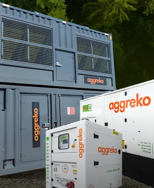 Three Aggreko power units, two white generators, and a large, stacked gray container unit, all on dirt in front of a forest.