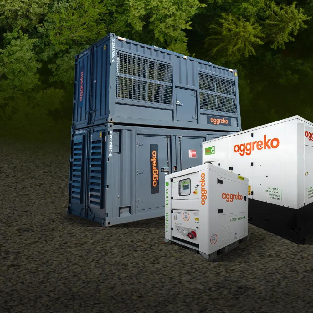 Three Aggreko power units, two white generators, and a large, stacked gray container unit, all on dirt in front of a forest.