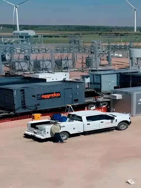 Aerial view of an industrial staging area next to an electrical substation. Several dark, containerized Aggreko industrial units are mounted on trailers on a gravel lot, with two white pickup trucks parked nearby in the dry, open landscape. Wind turbines visible in the distant background.