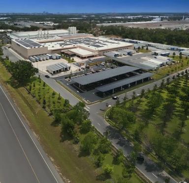 An aerial view of a large, single-story industrial or distribution center beside a road leading toward a city skyline.