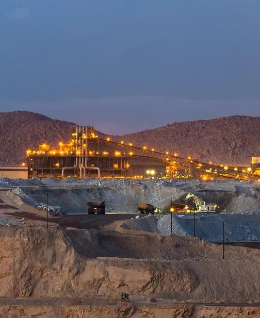 A mountainous landscape at dusk, featuring illuminated mining operations and snow-capped peaks in the background.