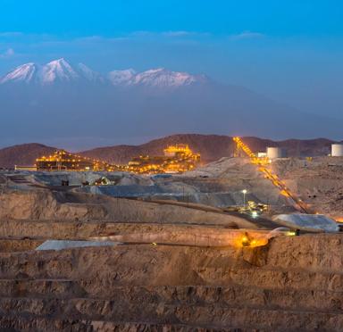 A mountainous landscape at dusk, featuring illuminated mining operations and snow-capped peaks in the background.