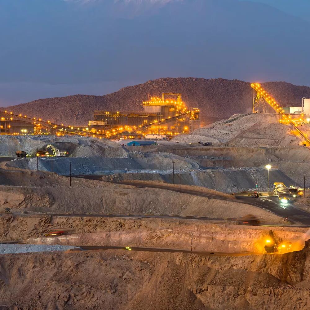 A mountainous landscape at dusk, featuring illuminated mining operations and snow-capped peaks in the background.