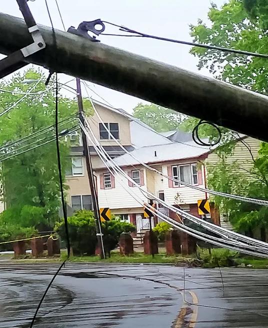 A downed utility pole with wires on a wet road, surrounded by greenery and a house in the background.