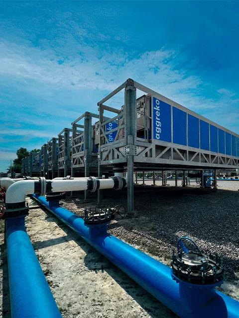 Vue en contre-plongée d’une longue unité industrielle bleue en conteneur, montée sur une haute structure métallique grise, avec plusieurs unités similaires s’estompant à l’arrière-plan sous un ciel nuageux.