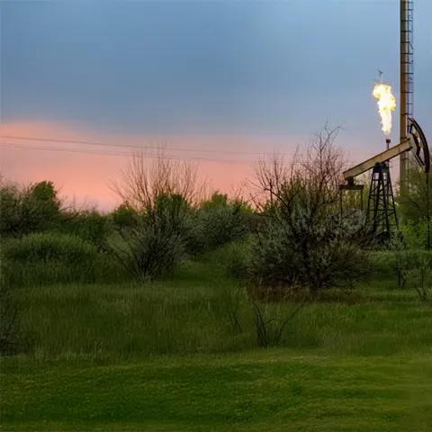 Une plateforme pétrolière avec une flamme qui brûle au sommet, entourée de verdure et d’un ciel nuageux.