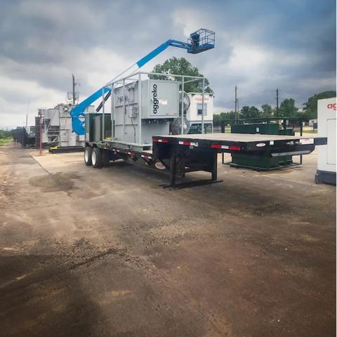 A modular, containerized Aggreko substation unit on the back of a flatbed trailer beside a green forest lining.