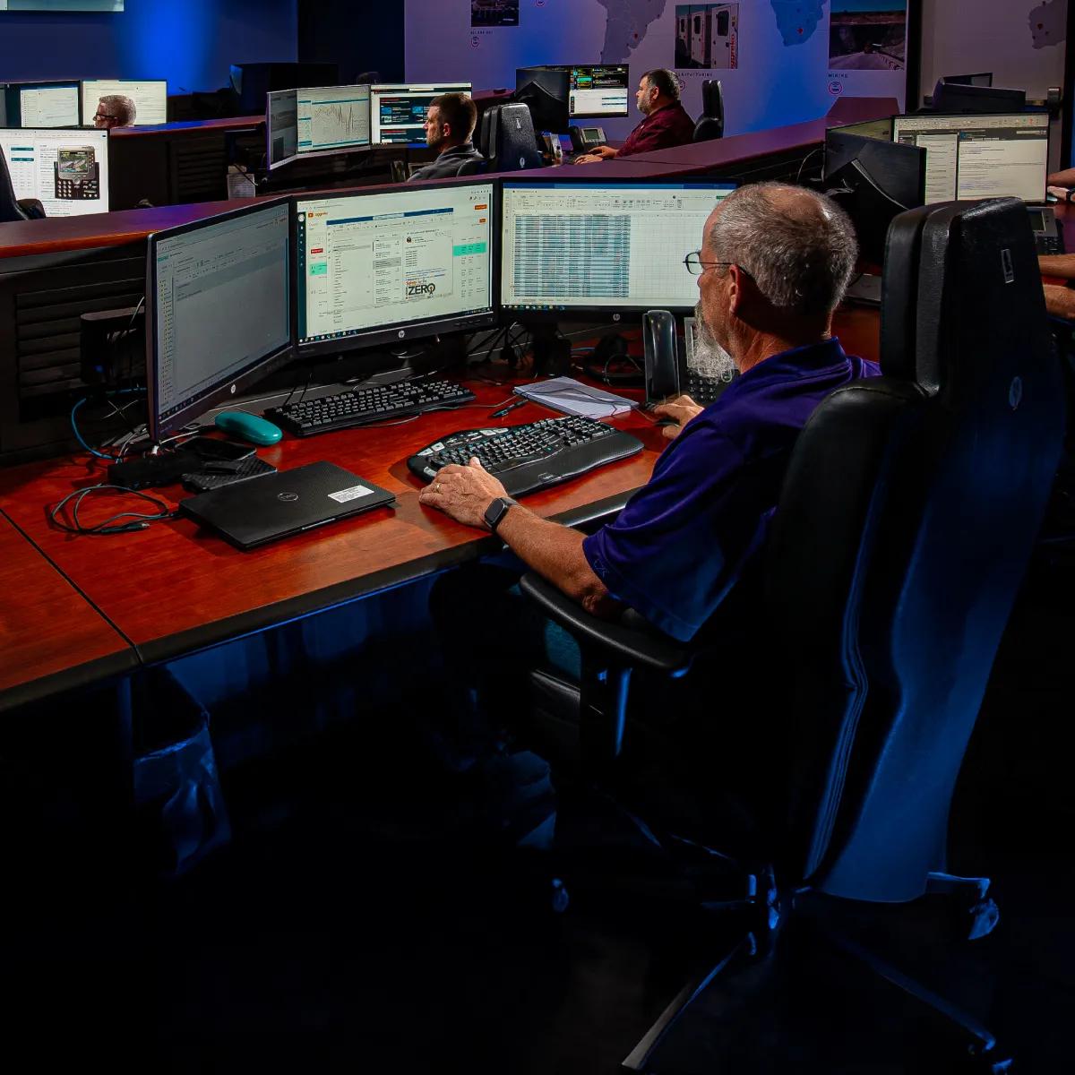 A dimly lit room with several people at workstations, focused on multiple computer screens displaying data and charts.