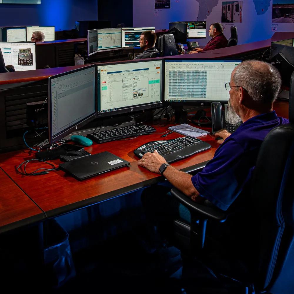 A person at a computer desk, working on multiple monitors in a busy control room environment.