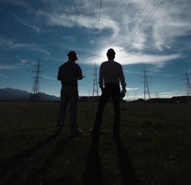 Shadow of two workers standing in a field seeing three main transmission towers and multiple towers in the background