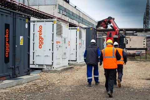 Three workers in uniform, walking by various white and black Aggreko containers, and towards a red crane.  