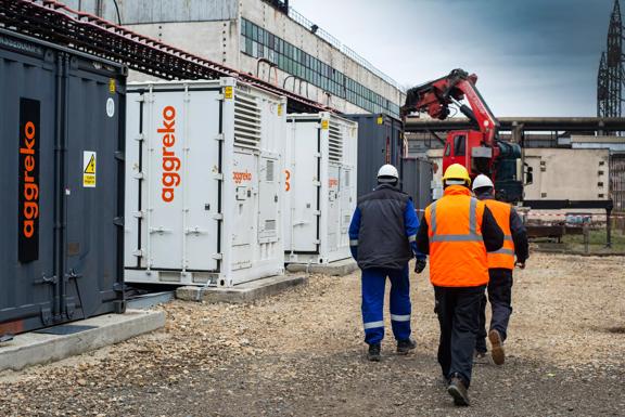Technicians in safety gear walking past Aggreko power units at an industrial worksite