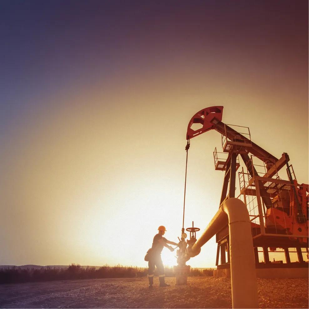 A worker next to an oil rig on a field against a sunset