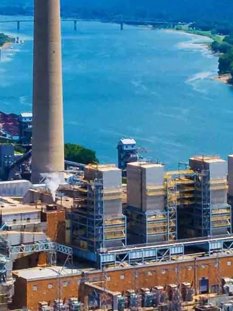 An aerial view of a large power plant and associated electrical substation next to a wide river and forested hills.