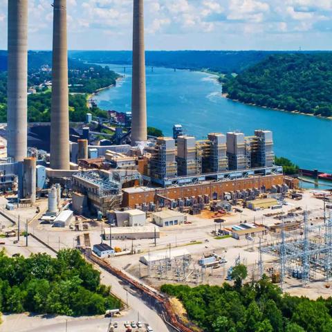 An aerial view of a large power plant and associated electrical substation next to a wide river and forested hills.