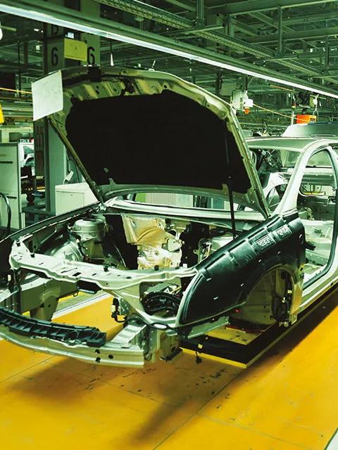 A brightly lit, close-up interior shot of an automotive assembly line, showing the metal frame, and open door and hood of a car body being constructed.