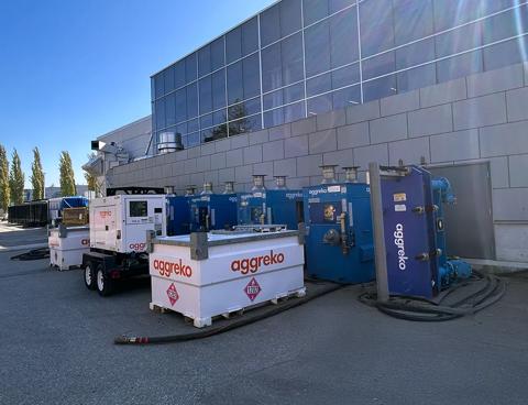 A bright, outdoor shot showing a row of large, modular industrial units positioned against the sleek, modern glass and metal facade of a commercial building.