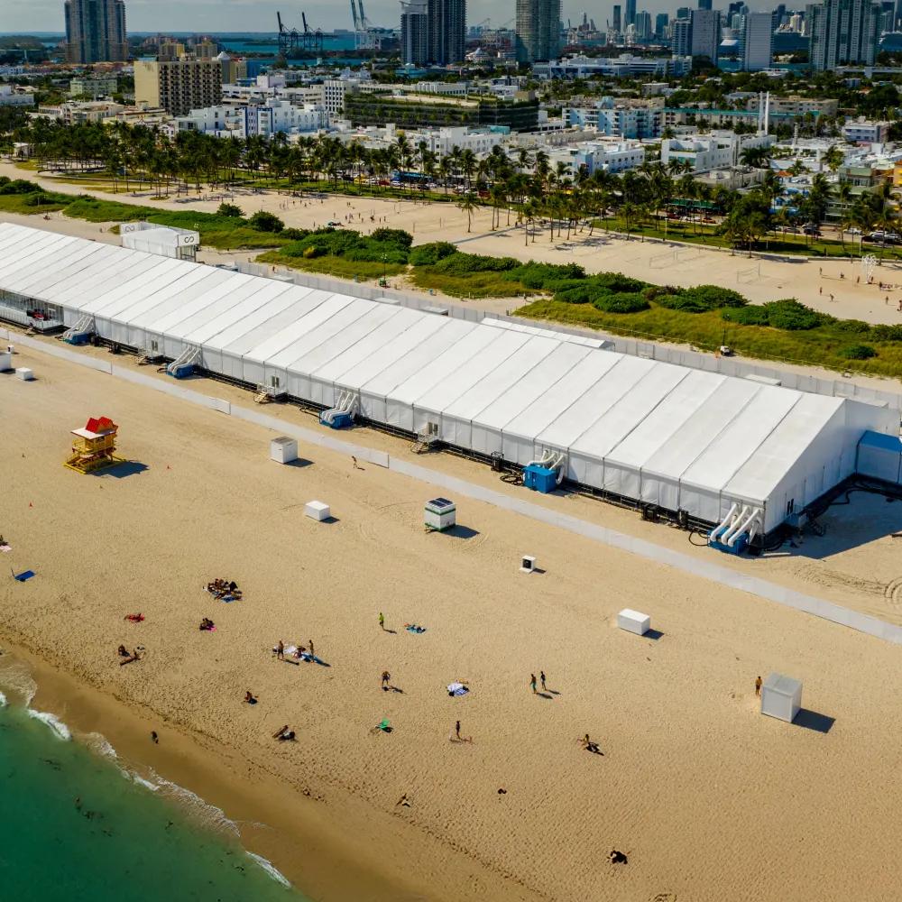 Aerial view of a big tent on the beach with people walking by and the city in the background.