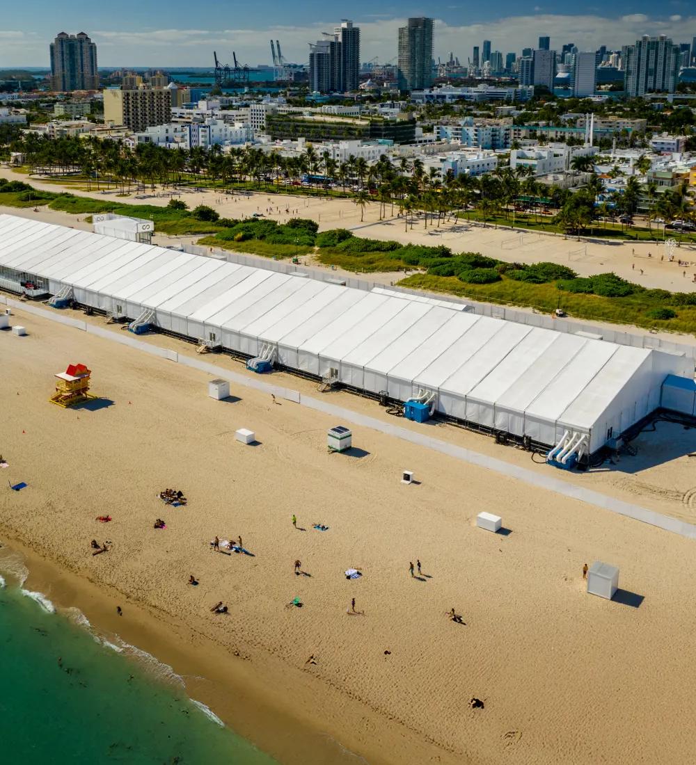 Aerial view of a big tent on the beach with people walking by and the city in the background.